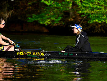A woman in a white visor sits on a rowboat facing a woman who is rowing.